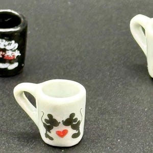 May include: Three miniature ceramic mugs. One mug is black with a white cartoon mouse character. Two mugs are white, one featuring a cartoon mouse couple with a red heart, and the other showing a cartoon mouse couple with red accents.