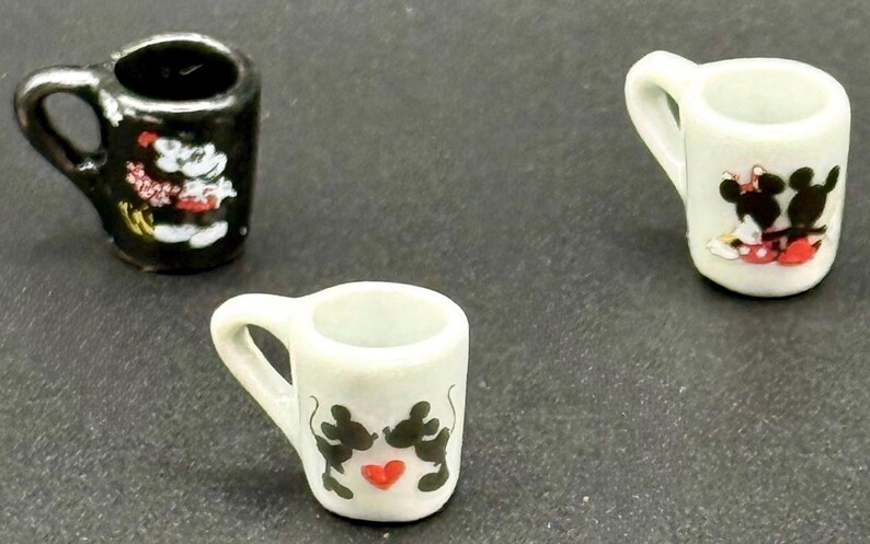 May include: Three miniature ceramic mugs. One mug is black with a white cartoon mouse character. Two mugs are white, one featuring a cartoon mouse couple with a red heart, and the other showing a cartoon mouse couple with red accents.
