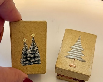 May include: Two small, rectangular, beige Christmas ornaments. One features two green Christmas trees with gold star toppers. The other has a silver and white tree design. Both ornaments have a gold border.