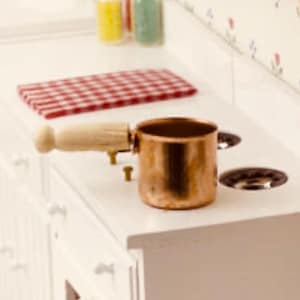 Puede incluir: Una pequeña cacerola de cobre con un mango de madera clara descansa sobre una superficie blanca. Un paño a cuadros rojo y blanco y pequeños frascos están en el fondo. La escena evoca una cocina.