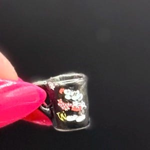 May include: A miniature black mug charm featuring a small white and red cartoon character graphic on the front. The tiny mug has a curved handle on the side and a smooth, glossy finish, designed to be used as a decorative accessory or jewelry component.