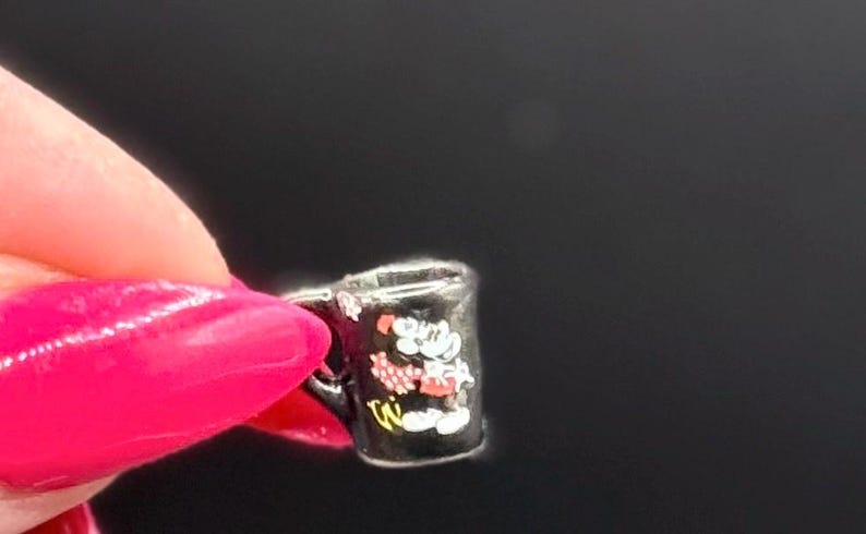 May include: A miniature black mug charm featuring a small white and red cartoon character graphic on the front. The tiny mug has a curved handle on the side and a smooth, glossy finish, designed to be used as a decorative accessory or jewelry component.