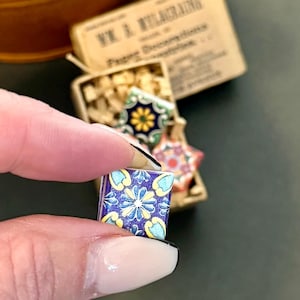 May include: A hand holds a small, square, decorative tile with a blue and yellow floral pattern. Other tiles with different patterns are in a small box. The box has the text "V. & A. WILBURNING" on it.