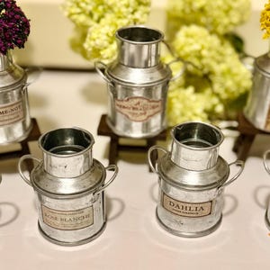 May include: A collection of small, galvanized metal milk can vases, each with a label and a handle. Some vases hold dried flowers in shades of purple and yellow. The vases are arranged on a white surface.