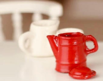 May include: A miniature red ceramic teapot with a matching lid, positioned in front of two white ceramic pitchers. The teapot has a handle and a spout. The background includes a white chair.