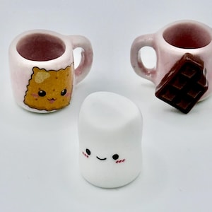 Może przedstawiać: Dwa miniaturowe kubki z jasnoróżowej ceramiki i figurka białej pianki marshmallow. Jeden kubek przedstawia żółtą, uśmiechniętą kwadratową postać, a drugi ma przymocowaną z boku brązową tabliczkę czekolady. Biała pianka ma małą buzię.