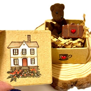 May include: A miniature scene featuring a brown teddy bear, a suitcase with a red heart, and a house illustration on a small box. The box is filled with paper and sits on a wooden slice. The house illustration has a red door and flower details.