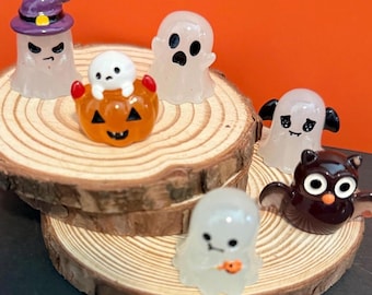 May include: A collection of Halloween-themed miniature figurines. Includes a ghost wearing a purple witch hat, several translucent ghosts, a jack-o'-lantern, and a brown owl. The figurines are arranged on a wooden slice against an orange background.