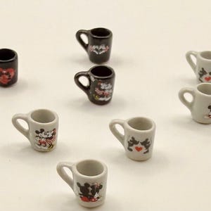 May include: A collection of ten miniature ceramic mugs in black and white, each featuring small graphic prints of cartoon characters and red heart motifs. The mugs are arranged in a scattered pattern, showcasing various designs on their surfaces.
