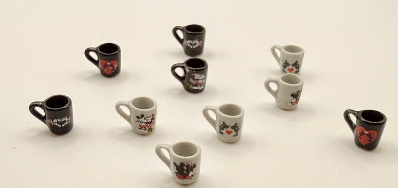 May include: A collection of ten miniature ceramic mugs in black and white, each featuring small graphic prints of cartoon characters and red heart motifs. The mugs are arranged in a scattered pattern, showcasing various designs on their surfaces.
