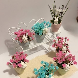 以下が含まれることがあります： ピンクとブルーの花と白いウサギの置物が入ったミニチュア鉢植え。植物は白い金属製のベンチと小さなテーブルに配置され、ベージュのプレースマットが敷かれています。春またはイースターをテーマにしたシーンです。