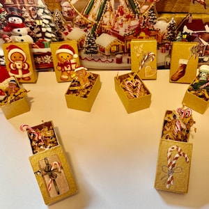 May include: A collection of small, gold-colored boxes, each with a Christmas-themed design. Some boxes contain candy canes and decorative filler, while others feature gingerbread figures and festive illustrations. The boxes are arranged on a white surface.