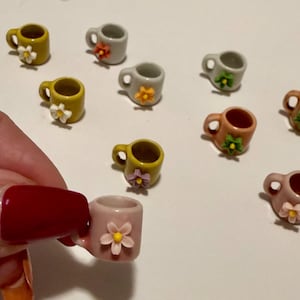 Peut inclure: Petites tasses en céramique de différentes couleurs, dont jaune, gris et rose, chacune ornée d'une petite fleur. Les tasses, avec une anse, sont disposées sur une surface blanche. Une tasse est tenue.
