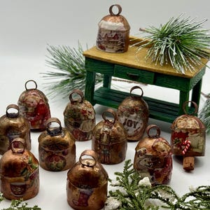 May include: A collection of twelve small, dome-shaped metal bells with a distressed copper finish, each featuring unique vintage-style holiday illustrations, including a nutcracker, a snowman, and the word JOY, arranged around a small green wooden table.