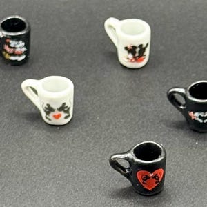 May include: A collection of miniature ceramic mugs in black and white, each featuring small graphic prints of hearts and cartoon characters. These tiny vessels have handles and are arranged in a scattered pattern across the frame.
