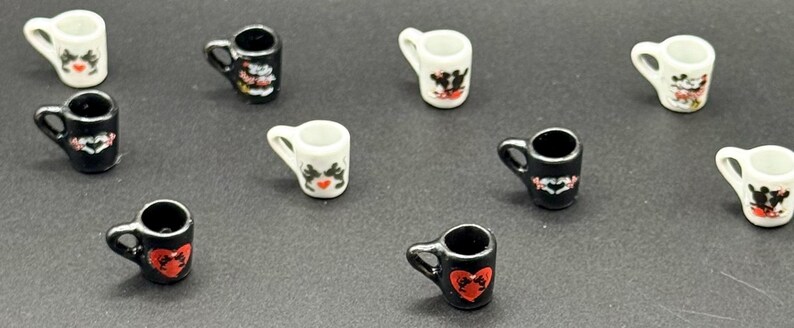 May include: A collection of miniature ceramic mugs in black and white, each featuring small graphic prints of hearts and cartoon characters. These tiny vessels have handles and are arranged in a scattered pattern across the frame.