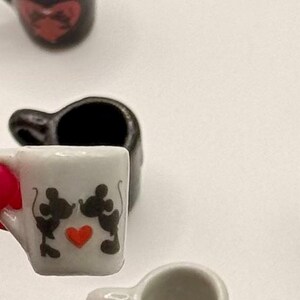 May include: A miniature white ceramic mug featuring a black silhouette graphic of two mice kissing with a small red heart in the center. The mug has a small handle on the side and is shown in a collection of similar tiny drinkware items.
