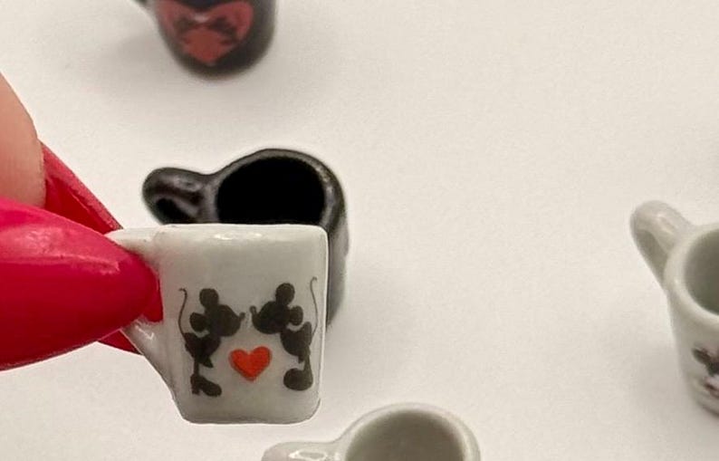 May include: A miniature white ceramic mug featuring a black silhouette graphic of two mice kissing with a small red heart in the center. The mug has a small handle on the side and is shown in a collection of similar tiny drinkware items.