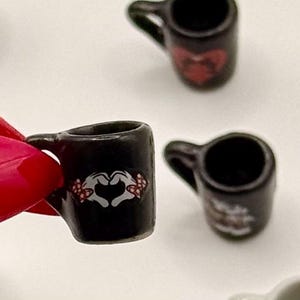 May include: A miniature black ceramic mug featuring a graphic of white cartoon hands forming a heart shape with red and white polka dot bows on the wrists. The small mug has a curved handle and a smooth, glossy finish, designed for dollhouse displays.