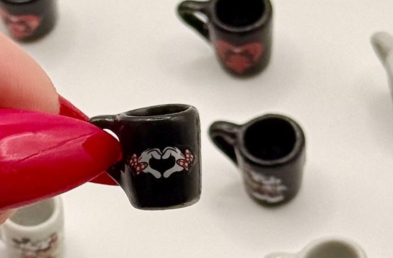 May include: A miniature black ceramic mug featuring a graphic of white cartoon hands forming a heart shape with red and white polka dot bows on the wrists. The small mug has a curved handle and a smooth, glossy finish, designed for dollhouse displays.