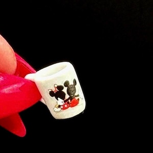 May include: A miniature white ceramic mug featuring a small graphic print of two cartoon mice characters, one in black and one in black with a red skirt, positioned against a plain black background to show the scale of the tiny drinkware item.