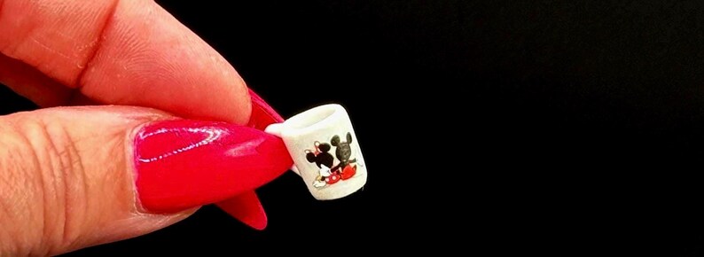 May include: A miniature white ceramic mug featuring a small graphic print of two cartoon mice characters, one in black and one in black with a red skirt, positioned against a plain black background to show the scale of the tiny drinkware item.