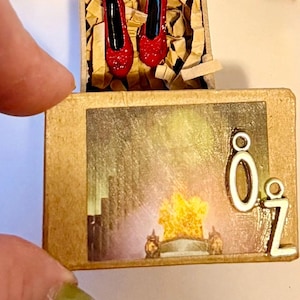 Può includere: Un diorama in legno in miniatura con una scena de Il Mago di Oz. Il diorama include una raffigurazione della Città di Smeraldo, le lettere "Oz" e un paio di scarpe rosse scintillanti. Le scarpe sono in una piccola scatola di legno con carta sminuzzata.