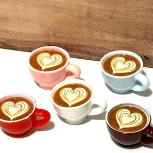 Pode incluir: Cinco pequenas chávenas de café, cada uma cheia de um líquido castanho e coberta com um desenho em forma de coração em espuma branca. As chávenas são de várias cores: vermelho, rosa, branco, azul claro e vermelho escuro. As chávenas estão dispostas numa superfície branca.