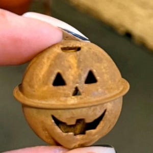 May include: A small, brown, pumpkin-shaped ornament with a smiling jack-o'-lantern face. The ornament has triangular eyes and a carved mouth. It is made of a material that appears to be metal or wood and is held between fingers.