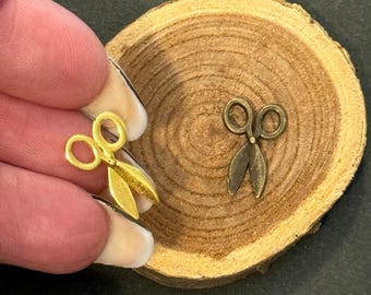 May include: Two small, decorative scissors charms. One is held by a hand and is gold-colored, while the other is a darker bronze color. The scissors are placed on a wooden slice, suggesting a craft or jewelry theme.