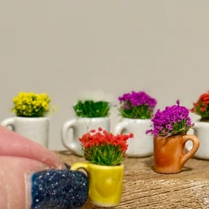 Op de afbeelding: Miniatuur keramische mokken gevuld met kleurrijke bloemen. De mokken zijn wit, geel en oranje, met bloemen in tinten geel, paars en rood. De mokken zijn gerangschikt op een houten oppervlak.