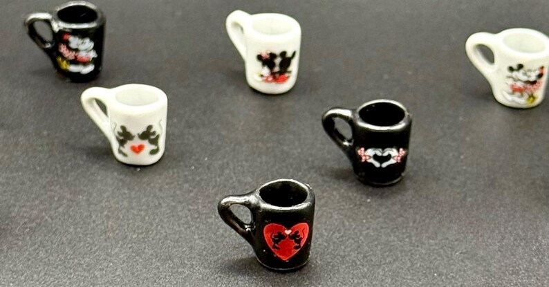 May include: A collection of miniature ceramic mugs in black and white, each featuring small graphic illustrations of cartoon mice. The mugs are arranged in a scattered pattern, showcasing various designs including heart motifs and silhouettes of mice.