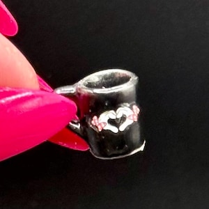 May include: A miniature black mug charm featuring a white graphic of two hands forming a heart shape. The design includes small pink accents on the hands. The mug has a silver-toned rim and a small handle on the side, presented as a tiny accessory.