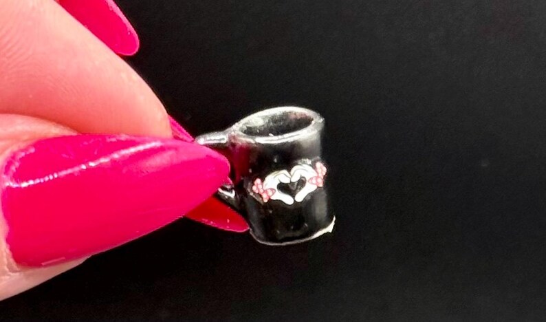 May include: A miniature black mug charm featuring a white graphic of two hands forming a heart shape. The design includes small pink accents on the hands. The mug has a silver-toned rim and a small handle on the side, presented as a tiny accessory.