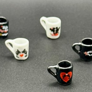 May include: A collection of nine miniature ceramic mugs in black and white. Each mug features a small handle and a decorative graphic on the front, including red heart shapes and black and white animal-like silhouettes, arranged in a scattered pattern.