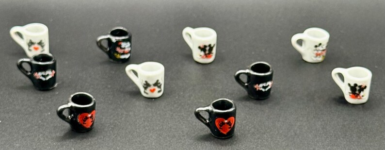 May include: A collection of nine miniature ceramic mugs in black and white. Each mug features a small handle and a decorative graphic on the front, including red heart shapes and black and white animal-like silhouettes, arranged in a scattered pattern.