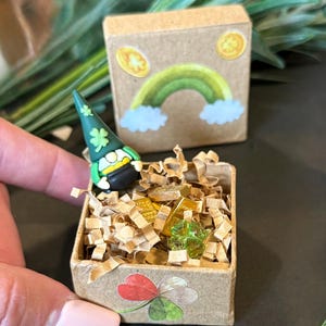 May include: A miniature St. Patrick's Day themed display featuring a small box filled with gold bars and coins, and a green shamrock. A second box has a rainbow and shamrock design. A gnome with a green hat is also present.