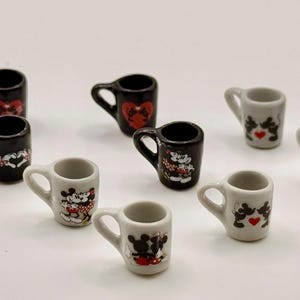 May include: A collection of nine miniature ceramic mugs in black and white. Each mug features a small handle and a graphic print of cartoon mice characters, some with red heart accents. The mugs are arranged in a scattered formation.
