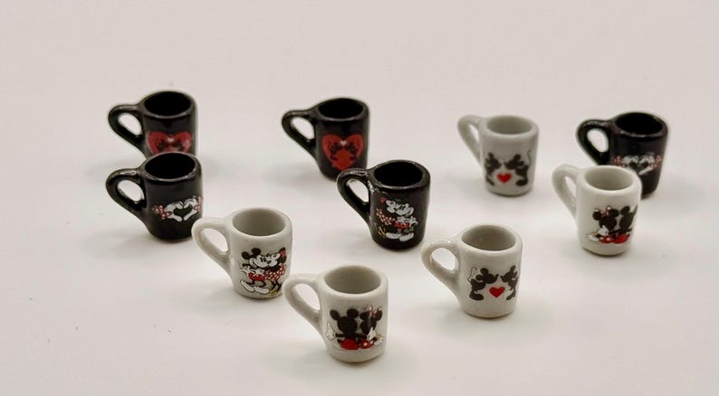 May include: A collection of nine miniature ceramic mugs in black and white. Each mug features a small handle and a graphic print of cartoon mice characters, some with red heart accents. The mugs are arranged in a scattered formation.