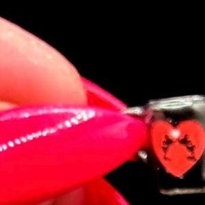 May include: A miniature black mug featuring a red heart graphic on the front. The small vessel has a cylindrical shape with a handle on the side and is held against a solid black background, showcasing its compact size and simple design.
