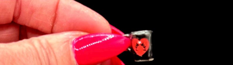 May include: A miniature black mug featuring a red heart graphic on the front. The small vessel has a cylindrical shape with a handle on the side and is held against a solid black background, showcasing its compact size and simple design.