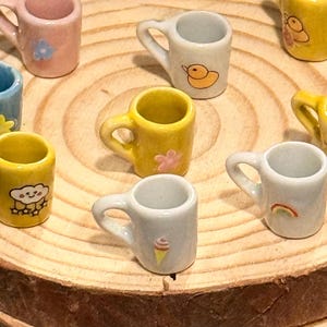 Pode incluir: Uma coleção de canecas de cerâmica em miniatura em várias cores, incluindo azul, amarelo, rosa e verde. Cada caneca apresenta um design diferente, como uma abelha, uma flor, um pato, uma nuvem e um arco-íris. As canecas estão dispostas numa superfície de madeira.