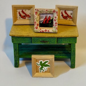 Op de afbeelding: Een kleine houten tafel met een groene basis en een gele bovenkant toont ingelijste kunst. Drie lijsten tonen rode schoenen en één heeft een groene fee. De tafel heeft een kleine lade.