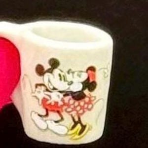 May include: A small white ceramic mug featuring a colorful graphic of Mickey Mouse and Minnie Mouse embracing. The mug has a simple cylindrical shape with a handle on the left side, showcasing the iconic cartoon characters in their classic attire.
