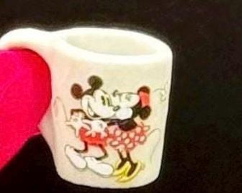 May include: A small white ceramic mug featuring a colorful graphic of Mickey Mouse and Minnie Mouse embracing. The mug has a simple cylindrical shape with a handle on the left side, showcasing the iconic cartoon characters in their classic attire.
