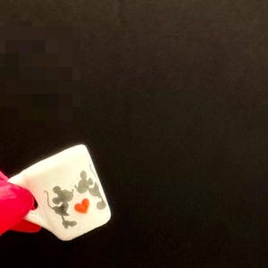 May include: A miniature white ceramic mug featuring a graphic of two grey mouse silhouettes facing each other with a small, vibrant red heart positioned between them. The mug has a small handle on the left side and a smooth, glossy finish.