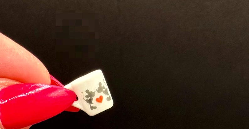 May include: A miniature white ceramic mug featuring a graphic of two grey mouse silhouettes facing each other with a small, vibrant red heart positioned between them. The mug has a small handle on the left side and a smooth, glossy finish.