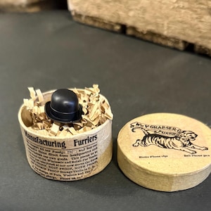 May include: A small black bowler hat rests in a cylindrical container filled with shredded paper. The container has printed text. A round box with a tiger illustration and text is next to it.