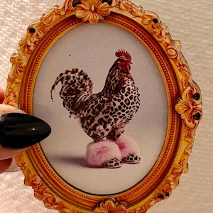 May include: An oval-shaped gold-toned ornate frame containing a print of a rooster with leopard-print feathers. The rooster is wearing fluffy pink slippers that also feature a leopard-print pattern on the toe area, set against a plain background.