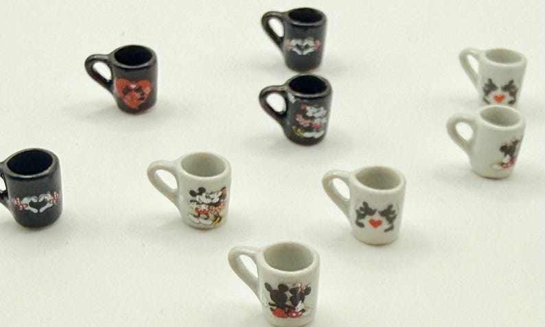 May include: A collection of miniature ceramic mugs in black and white, each featuring small graphic illustrations of cartoon characters and red heart accents. These tiny cups have a single handle and are arranged in a scattered pattern.